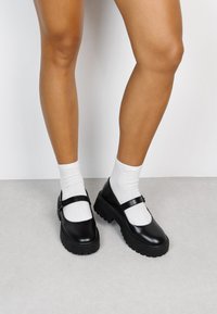 Zapatos Mary Jane negros con una suela de plataforma de goma gruesa y una tira con hebilla de metal, usados sobre calcetines tobilleros blancos. Textura suave.