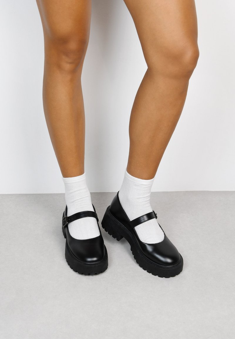 Zapatos Mary Jane negros con una suela de plataforma de goma gruesa y una tira con hebilla de metal, usados sobre calcetines tobilleros blancos. Textura suave.