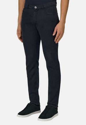 Mann trägt dunkelblaue Slim-Fit-Hose und schwarze Sneaker mit weißen Sohlen, steht vor einem schlichten hellen Hintergrund.