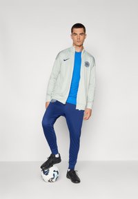 Lys grå lynlåsjakke med logo, blå skjorte og marineblå bukser. Sorte fodboldsko. Stående på en hvid og blå fodbold.