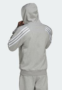 Felpa grigia con cappuccio e tasca a marsupio, caratterizzata da tre strisce bianche su ciascuna spalla. Realizzata in tessuto morbido, con vestibilità comoda e cappuccio regolabile.