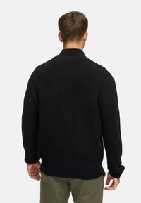 Zwarte gebreide trui met een hoge kraag, ribbelstructuur, verlaagde schouders en een relaxte pasvorm, gedragen over olijfgroene broeken.