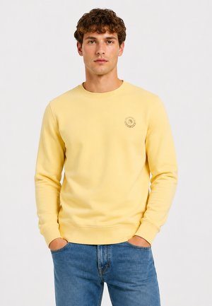 Jonge man met krullend haar, gekleed in een effen gele trui en blauwe spijkerbroek, staand met handen in de zakken tegen een witte achtergrond.