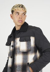 Podložena jakna s črnimi rokavi in kariranim telesom v beli, sivi in rjavi barvi. Ima velike žepne na prsih in gumbne zapiralo.