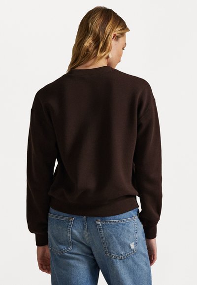 Brązowy bawełniany sweatshirt o luźnym kroju, okrągłym dekolcie i ściągaczach. Widoczny z tyłu, w połączeniu z niebieskimi dżinsami.