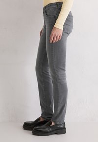 Personne portant un jean gris ajusté, des mocassins noirs et un haut à manches longues jaune pâle, adossée à un mur blanc texturé.