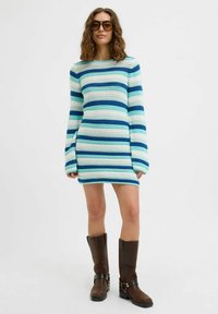 Prugasta pletena haljina u nijansama plave i zelene, s dugim rukavima i prilagođenim siluetom. Uparena s smešnim čizmama do koljena.