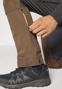Hand die zijrits van een bruine outdoor broek dicht ritsend over een grijze Columbia wandelschoen met een zwarte sok, tegen een effen achtergrond.