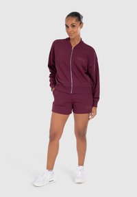 Sweat-shirt à zip bordeaux et shorts assortis, en tissu doux. Présente un logo sur la poitrine et sur les shorts, avec une coupe décontractée. Baskets blanches.