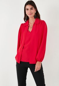 Red blouse with a pointed collar and buttons, featuring puffed sleeves and a flowy texture; styled over a v-neck top. Black pants worn.