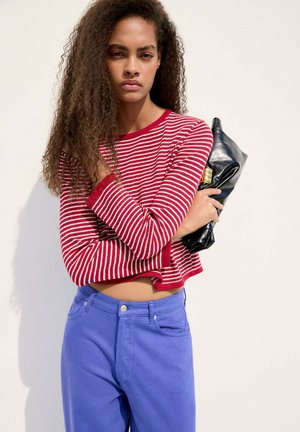Joven mujer con cabello largo y rizado, vistiendo una camiseta de rayas rojas y blancas y pantalones azules, sosteniendo un bolso de mano negro sobre un fondo liso.