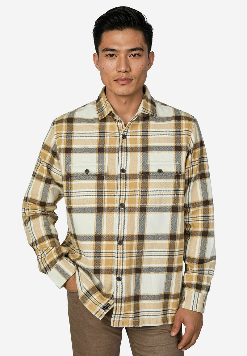 Mann trägt beige, braun und weiß kariertes Hemd mit Brusttaschen und braune Hose, steht mit einer Hand in der Tasche.