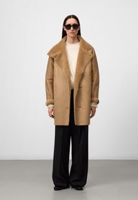 Vrouw in een oversized tan schapenvachtjas over een witte trui, zwarte brede broek, zwarte schoenen en zwarte zonnebril, staande tegen een effen achtergrond.