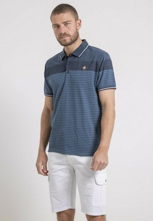 Homme aux cheveux courts et à la barbe, portant un polo à rayures bleues et un short cargo blanc, debout devant un fond uni.