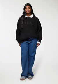 Felpa nera oversize con la scritta "AMOUR" in rilievo, camicia bianca con colletto sotto, jeans a gamba larga blu e sneaker grafiche trasparenti.