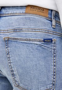 Vaqueros de mezclilla azul claro con costuras visibles, una etiqueta de cuero marrón que dice "PULL&BEAR" y una pequeña etiqueta azul en el bolsillo trasero.