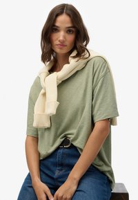 Olivengrøn T-shirt med løs pasform, parret med en cremefarvet strikket sweater bundet om halsen. Blå denimjeans og et sort bælte fuldender looket.