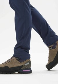 Bottes de randonnée en suède marron avec des semelles en caoutchouc noir et des accents violets ; associées à un pantalon ajusté bleu marine. Accent sur la conception et la texture des chaussures.