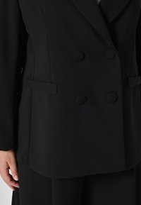 Nahaufnahme einer Person, die ein schwarzes doppelt geknöpftes Blazer mit mit Stoff überzogenen Knöpfen und vorderen Paspeltaschen trägt.