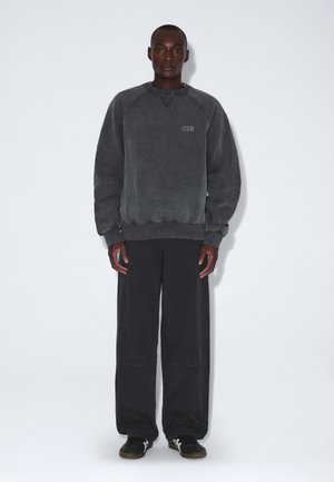 Mann, der vor einem einfarbigen Hintergrund steht, trägt ein ausgewaschenes dunkelgraues Sweatshirt, lockere schwarze Hose und schwarze Sneakers mit weißen Streifen.