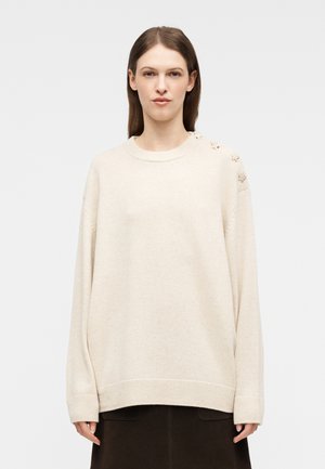 Mujer con cabello liso y castaño, vistiendo un suéter oversized color crema con botones en forma de estrella en el hombro izquierdo y una falda oscura.