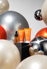 NIP+FAB-Produkte auf einem dunklen Sockel präsentiert, darunter eine orangefarbene Gel-Creme und ein Serum, umgeben von bunten, metallischen und matten Ballons.