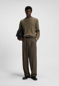 Khaki textured knit sweater, checkered brown trousers, black belt, and black shoes, with a black jacket draped over the arm.