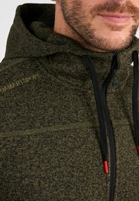 Dunkelgrüne Kapuzenjacke aus strukturiertem Strickstoff, mit schwarzem Reißverschluss und Kordeln mit roten Akzenten; enthält Logodetail am Kragen.