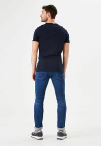 Marineblauw, nauwsluitend shirt met korte mouwen. Draagt een lichtblauwe spijkerbroek met opgerolde zomen. Grijze en marineblauwe sportschoenen zichtbaar.