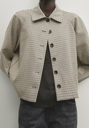 Person trägt eine beige-braune, kleine karierte Knopfjacke über einem dunklen Hemd, mit den Händen in den Taschen, vor einem einfarbigen hellen Hintergrund.