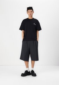 Tricou negru cu mânecă scurtă și logo alb, combinat cu pantaloni scurți gri până la genunchi și pantofi negri masivi. Modelul stă pe un fundal neutru.