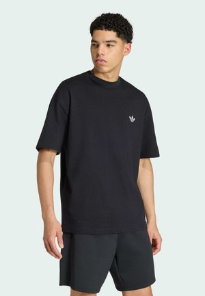 Junger Mann trägt ein schwarzes Adidas T-Shirt mit Logo auf der Brust und schwarze Shorts, steht vor einem schlichten hellen Hintergrund und schaut nach links.