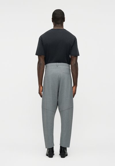 Ανδρας που φοράει μαύρο κοντομάνικο πουκάμισο, γκρι ψηλόμεσα παντελόνια με ραφές και μαύρα μποτάκια αστραγάλου, στέκεται με την πλάτη γυρισμένη σε απλό φόντο.