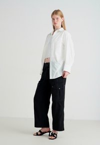 Jonge vrouw in een oversized wit overhemd, zwarte cargobroek en zwarte open teen hakken, staande tegen een eenvoudige witte achtergrond.