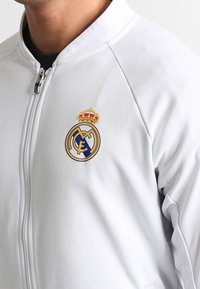 Witte zip-up jas gemaakt van gladde stof. Kenmerkt zich door een gedetailleerd geborduurd Real Madrid-logo met een gouden kroon en blauwe accenten.