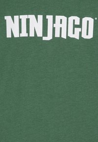 Green cotton t-shirt featuring large white text reading “NINJAGO.” Smooth texture with a round neckline and short sleeves.