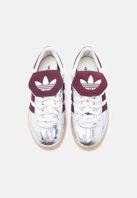 Ζευγάρι ασημί μεταλλικά αθλητικά παπούτσια Adidas με μπορντό ρίγες, κορδόνια και μεγάλα λογότυπα από πάνω, απεικονιζόμενα από πάνω σε λευκό φόντο.