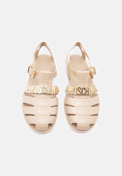 Sandales en gel beige Moschino avec détail de logo doré sur les brides et fermetures à boucle ajustables en or, vues de dessus sur fond blanc.