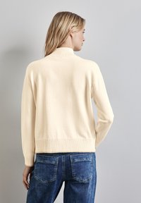 Pullover beige chiaro con collo alto e polsini a costine, caratterizzato da una superficie liscia e spacchi laterali, abbinato a jeans blu scuro.