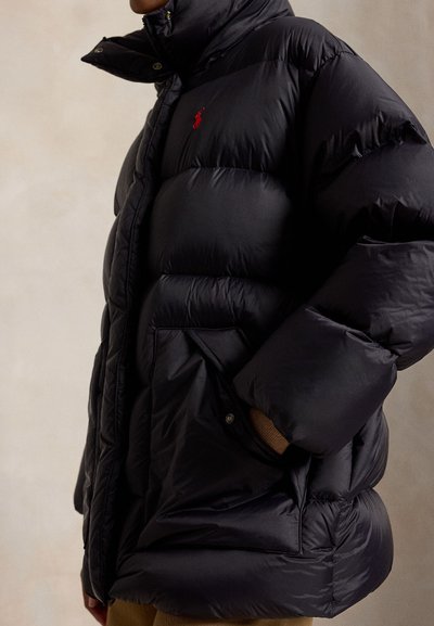 Person iført en sort dunjakke med et rødt logo og hænderne i forlommerne mod en ensfarvet baggrund.