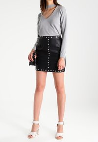 Vrouw in grijze V-hals lange mouw top met choker detail, zwarte leren minirok met studs, witte hoge hakken en een zwarte schoudertas met ketting.
