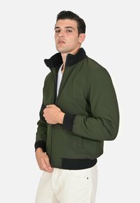 Giacca bomber verde oliva con colletto e polsini neri, con zip frontale e tasche laterali, realizzata in un tessuto liscio e testurizzato.