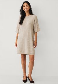 Beiges Strickkleid mit kurzen Ärmeln, lockerer Passform, Rundhalsausschnitt und geripptem Saum. Getragen mit dunkelbraunen spitzen Pumps.