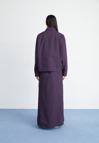 Violetti, kaksiosainen asu, joka koostuu pitkistä hameesta ja kauluksettomasta takista. Kangas on sileä, ja hameessa on selvästi näkyvät sivutaskut.