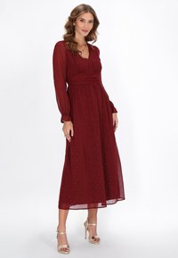 Rochie Bordeaux cu mânecă lungă, cu un corsaj ajustat, fustă evazată și un model brodat delicat. Asortată cu sandale cu toc cu curea aurii.