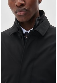 Cappotto nero con colletto alto, chiusura con zip e texture liscia. Indossato sopra una camicia a righe e una cravatta. Nessun motivo visibile o decorazioni.