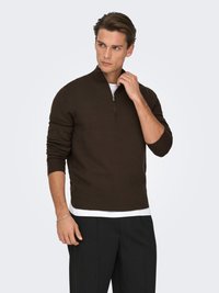 Brown zip-up sweater with a high collar, ribbed cuffs, and a fitted design, worn over a white shirt and paired with dark trousers.