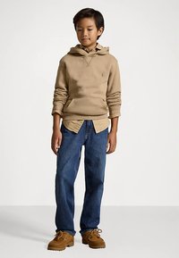 Μπέιζ ζακέτα με τσέπη καγκουρό, μπλε τζιν και καφέ δετά παπούτσια. Η ζακέτα διαθέτει ένα μικρό λογότυπο στο στήθος.