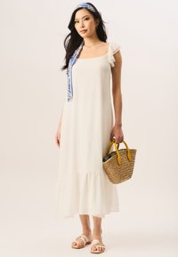 White maxi dress with ruffled sleeves, tiered hem, paired with a woven straw bag featuring yellow handles and flat sandals. Blue patterned scarf accent.