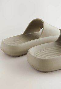 Bežiniai guminiai sandalai su plačiu dirželiu ir tekstūruotu padu. Yra apvalios, atviros nosies dizainas ir storas, paminkštintas padas.